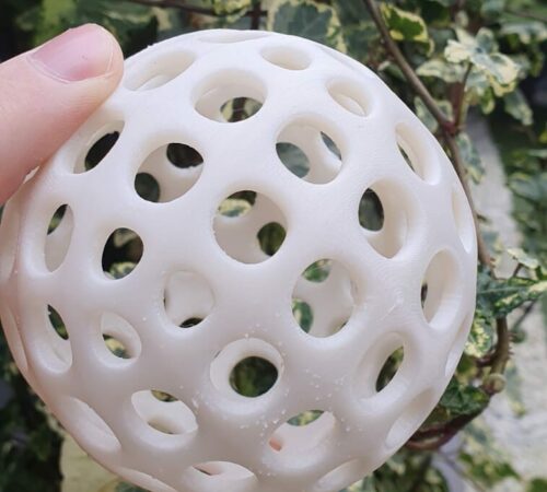 White perforated 3D-printed sphere held in a hand against green foliage.